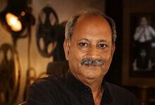 A man with a moustache and grey hair, wearing a black shirt, smiling in a warmly lit room with vintage de.