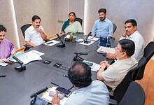 A woman and five men in a business meeting around a conference table with laptops and documents.