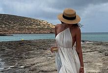 Relaxed woman in a white dress and straw hat holding a drink on a rocky beach with a scenic ocean view.