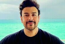 A man with dark hair and a beard smiling at the camera on a beach with turquoise water and a cloudy sky.