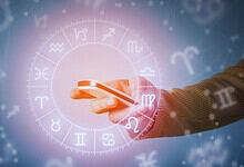 Hand holding smartphone with zodiac wheel and astrological symbols in the background.