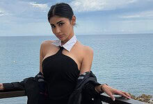 A woman in a stylish black dress and white scarf posing by the sea on a cloudy day.