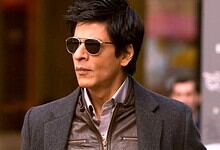 A woman with short dark hair wearing sunglasses and a grey blazer, standing outdoors in an urban setting.
