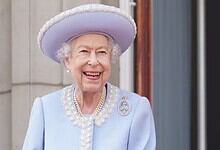 Queen Elizabeth II smiling in a light blue dress and hat, standing outdoors with a building in the背景.