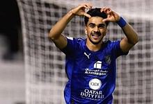 Football player celebrating goal with hands on head, smiling, wearing blue kit on the field.