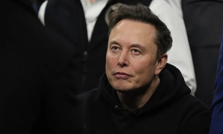 Elon Musk speaking at a public event, wearing a black hoodie, with a focused expression on his face.