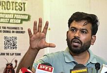 Man speaking at a press conference with protest poster in background.