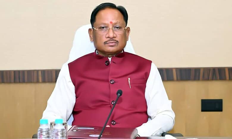 A senior Indian official wearing a maroon waistcoat and white shirt, seated at a desk with microphones an.