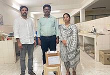 A woman and two men standing in a kitchen or food preparation area, with boxes and supplies around them.