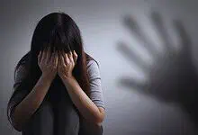 A woman with long dark hair covering her face, appearing distressed or upset, in a dimly lit room.