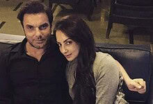 A man and woman sitting together on a black sofa in a dimly lit indoor setting.