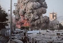 Explosion with thick black smoke rising over a cityscape with tents and buildings.