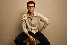 Tom Holland sitting on a stool, wearing a beige shirt and black pants, against a plain background.