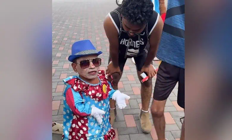 Child dressed as a clown with sunglasses and a blue hat, standing outdoors on a brick pavement.