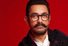 Aamir Khan with glasses and a moustache against a red background.