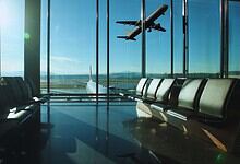 Empty airport departure lounge with rows of seating overlooking a runway as a plane takes off into a clear blue sky