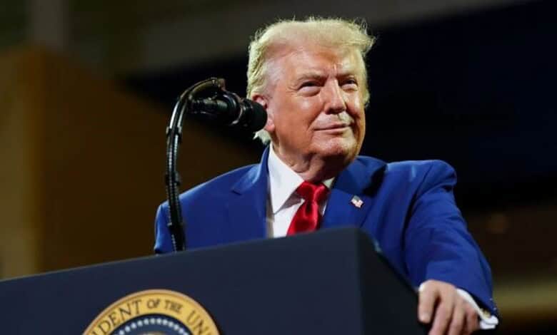 Donald Trump at a podium during a speech, wearing a blue suit and red tie, with a serious expression.