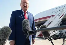 Donald Trump speaking at an airport with microphones in front, aircraft in background.