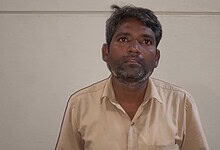 Man with a serious expression in beige shirt against plain wall background.
