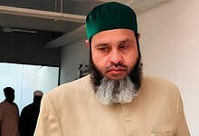 Close-up of a man with a beard wearing a green cap and beige traditional attire.