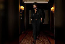 Nagarjuna in black suit walking confidently in a luxurious corridor.