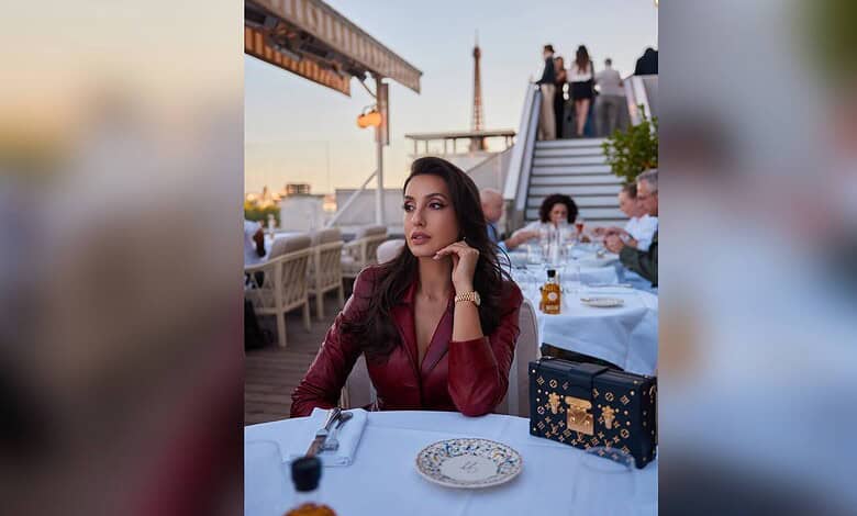 Nora Fatehi seated at a rooftop restaurant during sunset, with a city skyline in the background.