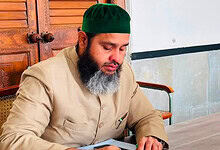 Man with a beard wearing a green cap and beige traditional attire sitting at a wooden table.