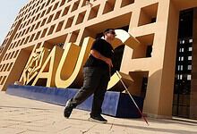 Blind Egyptian man with white cane walking past a modern building with large yellow letters.