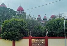 Historical building with red domes and lush greenery in front, under a cloudy sky.
