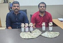 Two men seated at a table with confiscated drugs and cash displayed in front of them.