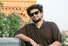 Young man with sunglasses and beard smiling outdoors in front of a monument and greenery.