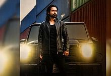 Man with long hair and beard standing in front of a black SUV at sunset.