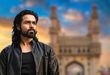 A man with long black hair and beard stands in front of a historic monument at sunset.