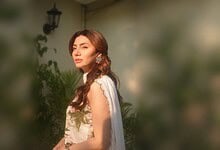 Mahira Khan with long wavy hair, wearing a white dress, standing outdoors in sunlight.