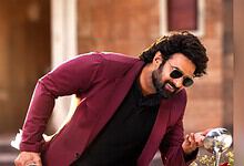Prabhas in a maroon blazer and sunglasses, smiling and looking at his phone outdoors.