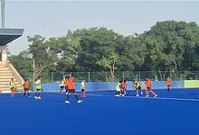 Young athletes training on a blue outdoor hockey field with coaches and lush green trees in the backdrop.