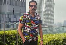 Man in colourful shirt and sunglasses standing on rooftop with cityscape background.
