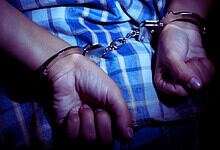 Person in handcuffs wearing a blue checkered shirt, close-up shot.