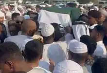 Large crowd of people attending a funeral procession outdoors in a sunny setting.