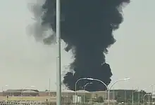 Thick black smoke rising from a fire near Riyadh airport, with clear sky and streetlights in the scene.