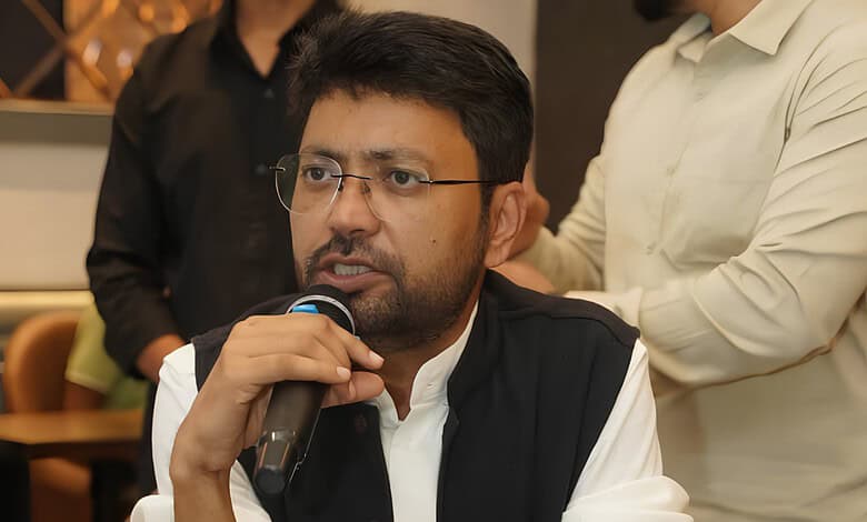 Man speaking into a microphone at a formal event, wearing glasses and a black vest.