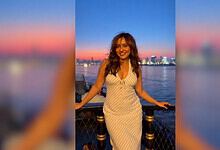 Neha Sharma smiling by the river during sunset with city skyline in background.