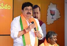 Telangana BJP chief speaking at a political event with party symbols and leaders in the background.