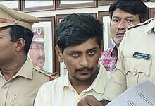 Young man with dark hair and mustache in police uniform, surrounded by officers, reading a document.