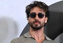 Man with curly hair, beard, sunglasses, wearing a grey shirt, against a plain background.