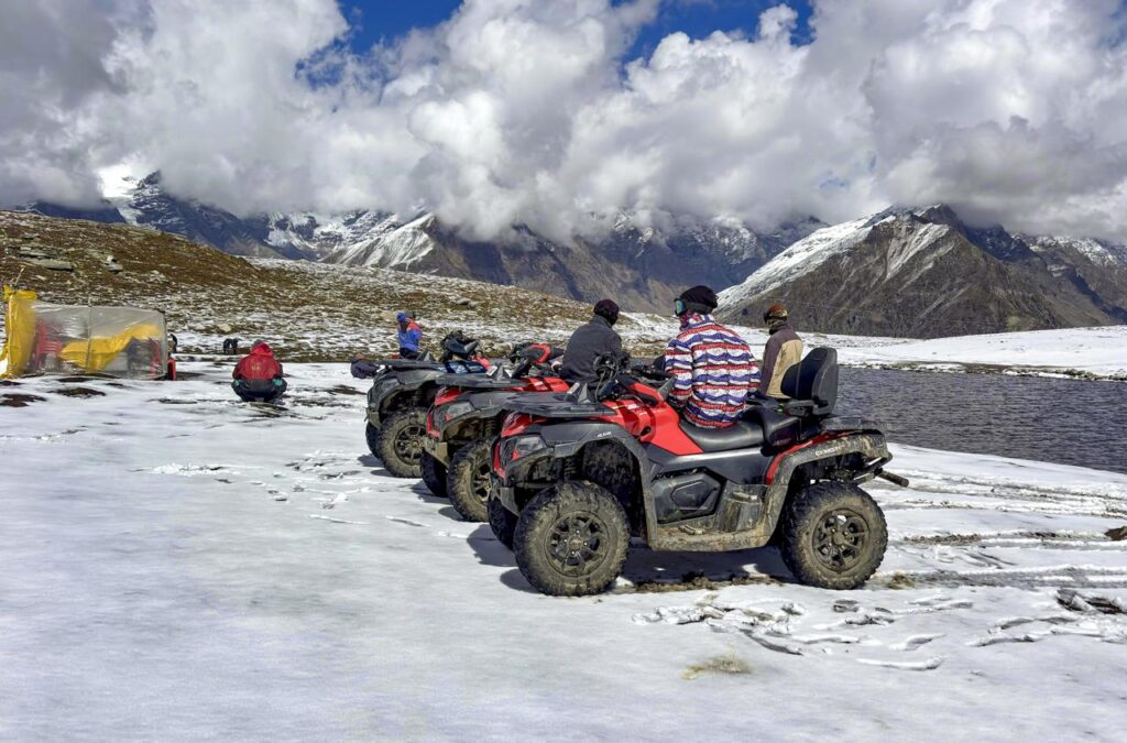 Tourists riding quad bikes on snow-covered landscape in Kashmir with mountains and clouds in background.
