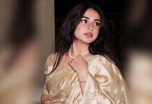Yumna Zaidi with dark hair, wearing a beige dress and pearl necklace, looking emotional on a flight.