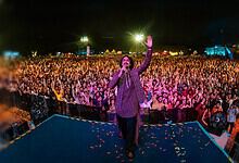 A speaker addresses a large crowd at night during a public event with bright lights and tents in the背景.