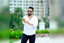Mika Singh wearing sunglasses and a white shirt, standing outdoors with a cityscape background.
