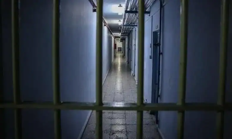 Empty prison corridor with barred cells and dim lighting.